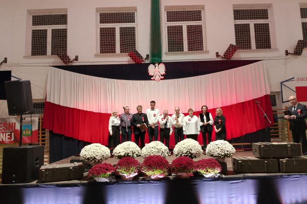 Zespół śpiewający na scenie z biało-czerwoną dekoracją i kwiatami podczas Festiwalu Pieśni Patriotycznych.