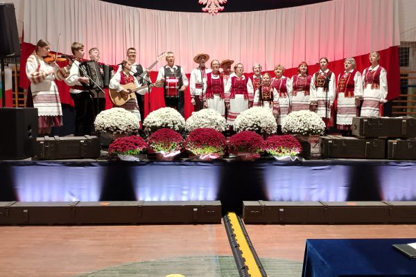 Zespół śpiewający na scenie z biało-czerwoną dekoracją i kwiatami podczas Festiwalu Pieśni Patriotycznych.