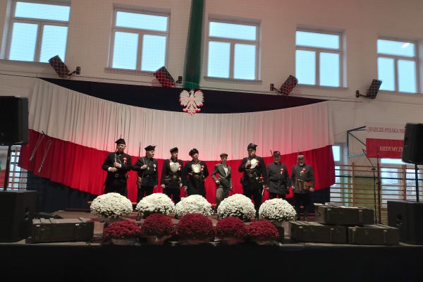 Zespół śpiewający na scenie z biało-czerwoną dekoracją i kwiatami podczas Festiwalu Pieśni Patriotycznych.