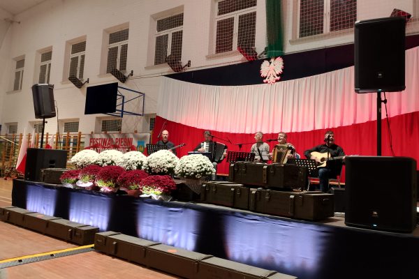 Zespół śpiewający na scenie z biało-czerwoną dekoracją i kwiatami podczas Festiwalu Pieśni Patriotycznych.