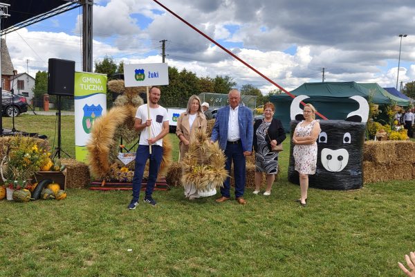 Grupa uczestników z wieńcem dożynkowym przed udekorowaną sceną.