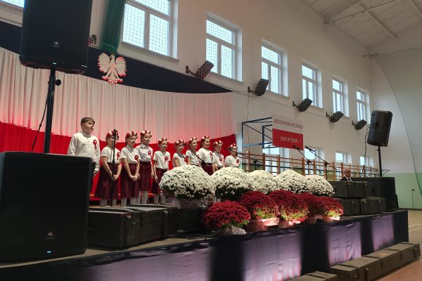 Zespół śpiewający na scenie z biało-czerwoną dekoracją i kwiatami podczas Festiwalu Pieśni Patriotycznych.