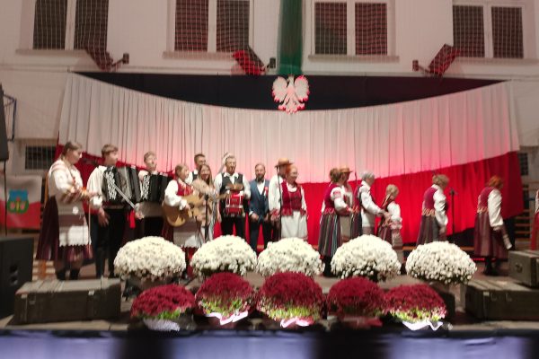 Zespół śpiewający na scenie z biało-czerwoną dekoracją i kwiatami podczas Festiwalu Pieśni Patriotycznych.