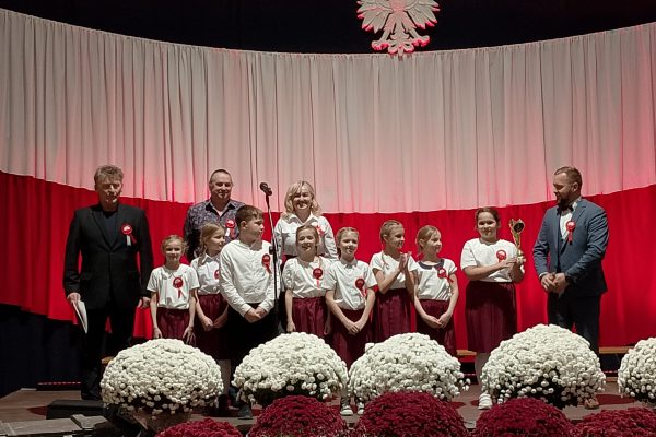 Zespół śpiewający na scenie z biało-czerwoną dekoracją i kwiatami podczas Festiwalu Pieśni Patriotycznych.