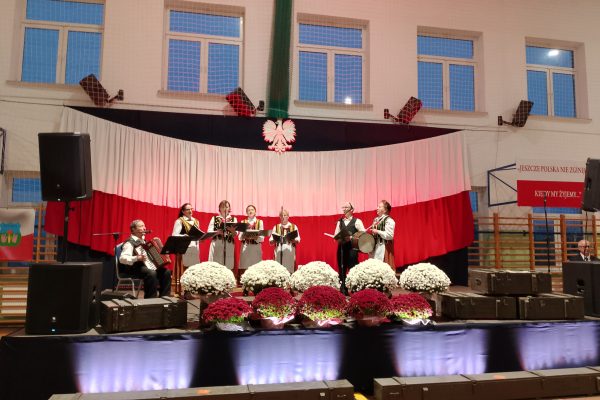 Zespół śpiewający na scenie z biało-czerwoną dekoracją i kwiatami podczas Festiwalu Pieśni Patriotycznych.