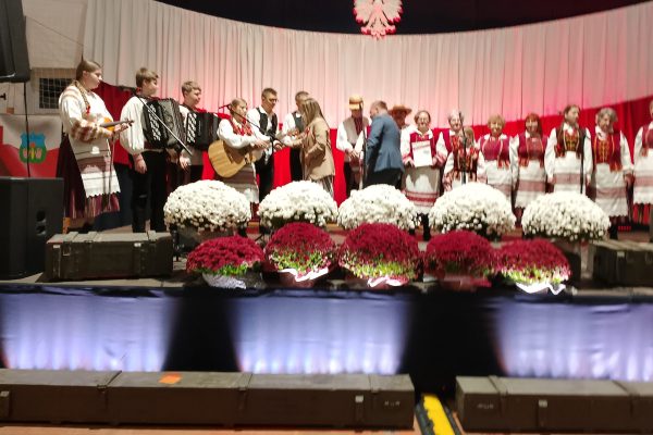Zespół śpiewający na scenie z biało-czerwoną dekoracją i kwiatami podczas Festiwalu Pieśni Patriotycznych.