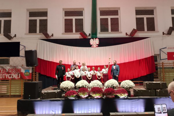 Zespół śpiewający na scenie z biało-czerwoną dekoracją i kwiatami podczas Festiwalu Pieśni Patriotycznych.
