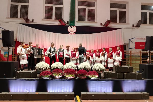 Zespół śpiewający na scenie z biało-czerwoną dekoracją i kwiatami podczas Festiwalu Pieśni Patriotycznych.