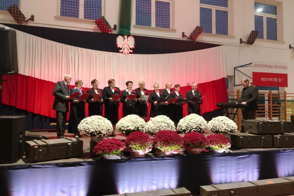 Zespół śpiewający na scenie z biało-czerwoną dekoracją i kwiatami podczas Festiwalu Pieśni Patriotycznych.