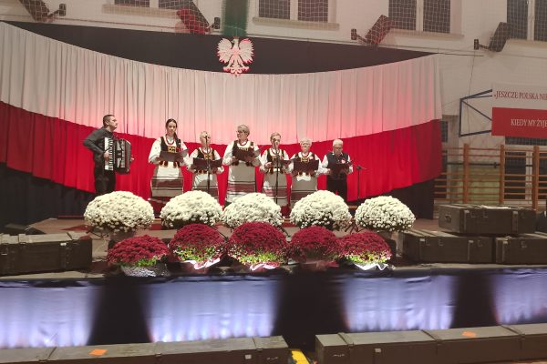 Zespół śpiewający na scenie z biało-czerwoną dekoracją i kwiatami podczas Festiwalu Pieśni Patriotycznych.