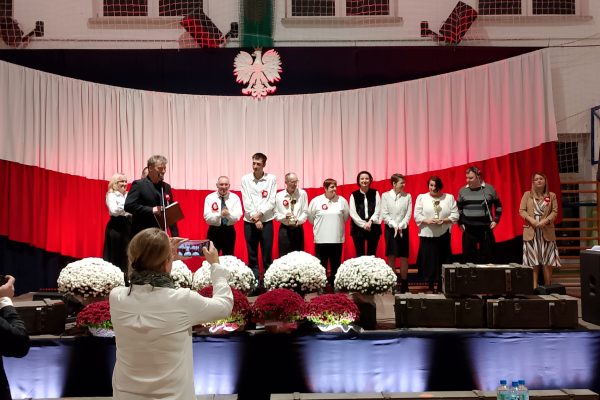 Zespół śpiewający na scenie z biało-czerwoną dekoracją i kwiatami podczas Festiwalu Pieśni Patriotycznych.