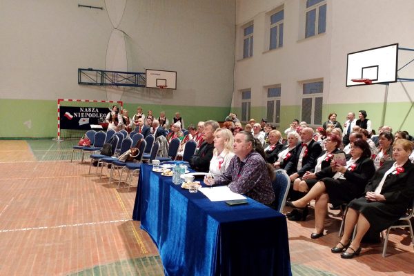 Publiczność i osoby przy stole jury podczas Festiwalu Pieśni Patriotycznych w sali sportowej.