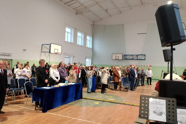 Publiczność i osoby przy stole jury podczas Festiwalu Pieśni Patriotycznych w sali sportowej.