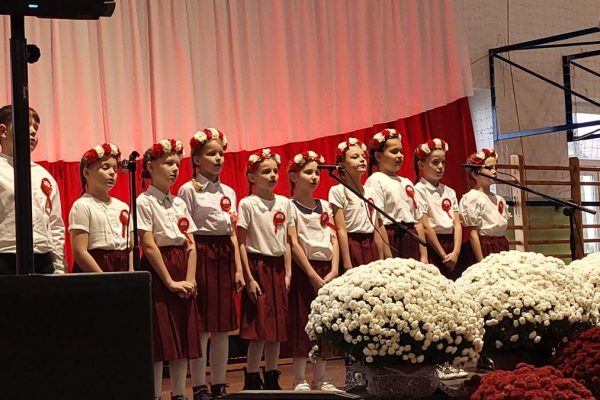 Zespół śpiewający na scenie z biało-czerwoną dekoracją i kwiatami podczas Festiwalu Pieśni Patriotycznych.