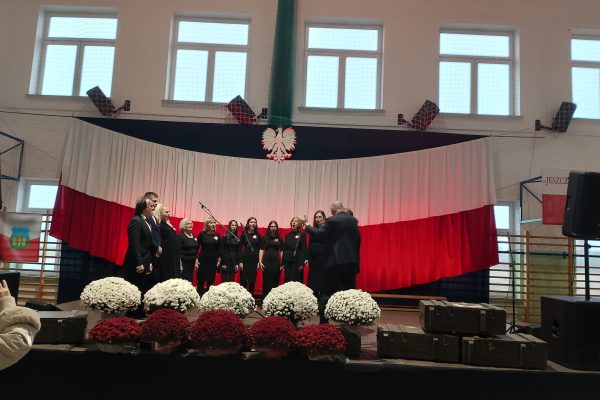 Zespół śpiewający na scenie z biało-czerwoną dekoracją i kwiatami podczas Festiwalu Pieśni Patriotycznych.