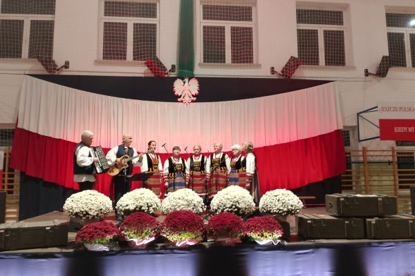 Zespół śpiewający na scenie z biało-czerwoną dekoracją i kwiatami podczas Festiwalu Pieśni Patriotycznych.