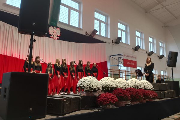 Zespół śpiewający na scenie z biało-czerwoną dekoracją i kwiatami podczas Festiwalu Pieśni Patriotycznych.