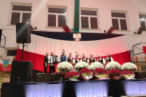 Zespół śpiewający na scenie z biało-czerwoną dekoracją i kwiatami podczas Festiwalu Pieśni Patriotycznych.