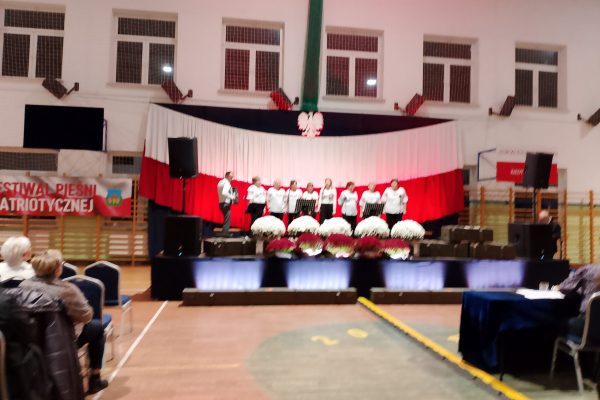 Zespół śpiewający na scenie z biało-czerwoną dekoracją i kwiatami podczas Festiwalu Pieśni Patriotycznych.