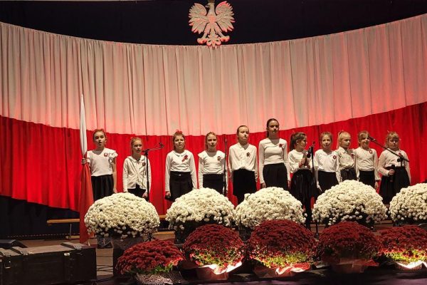 Zespół śpiewający na scenie z biało-czerwoną dekoracją i kwiatami podczas Festiwalu Pieśni Patriotycznych.