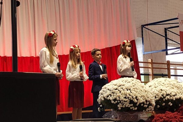 Zespół śpiewający na scenie z biało-czerwoną dekoracją i kwiatami podczas Festiwalu Pieśni Patriotycznych.