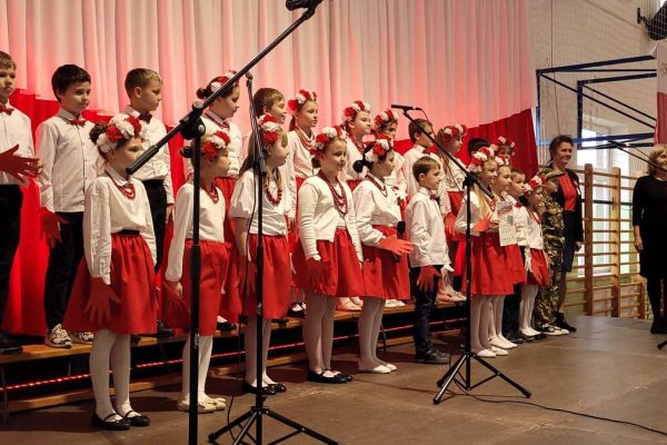 Zespół śpiewający na scenie z biało-czerwoną dekoracją i kwiatami podczas Festiwalu Pieśni Patriotycznych.