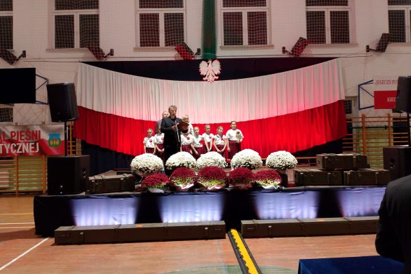 Zespół śpiewający na scenie z biało-czerwoną dekoracją i kwiatami podczas Festiwalu Pieśni Patriotycznych.
