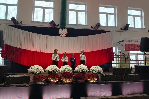 Zespół śpiewający na scenie z biało-czerwoną dekoracją i kwiatami podczas Festiwalu Pieśni Patriotycznych.