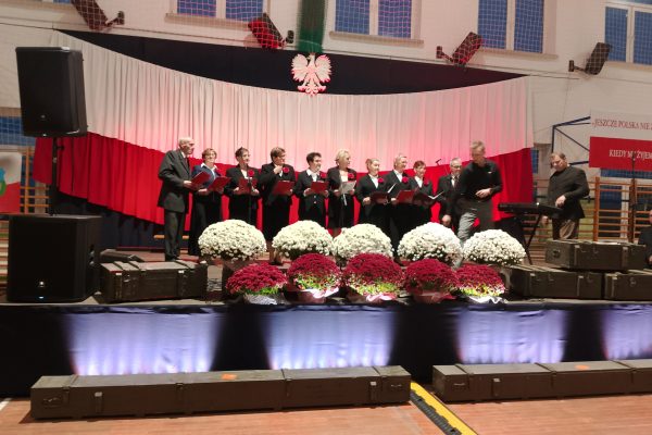 Zespół śpiewający na scenie z biało-czerwoną dekoracją i kwiatami podczas Festiwalu Pieśni Patriotycznych.