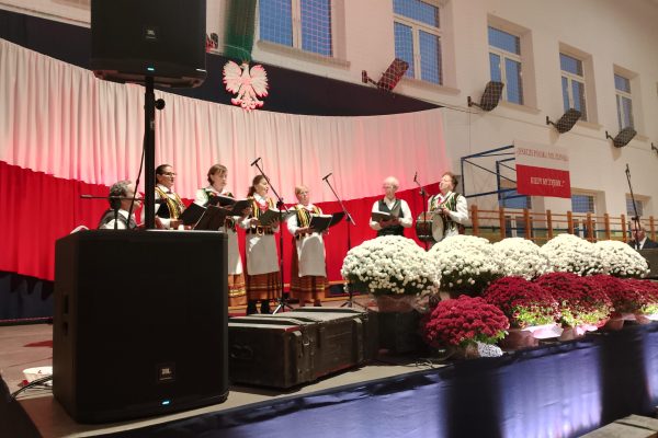 Zespół śpiewający na scenie z biało-czerwoną dekoracją i kwiatami podczas Festiwalu Pieśni Patriotycznych.