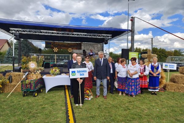Grupa uczestników z wieńcem dożynkowym przed udekorowaną sceną.