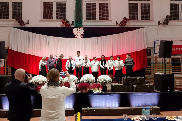 Zespół śpiewający na scenie z biało-czerwoną dekoracją i kwiatami podczas Festiwalu Pieśni Patriotycznych.