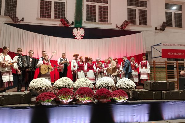 Zespół śpiewający na scenie z biało-czerwoną dekoracją i kwiatami podczas Festiwalu Pieśni Patriotycznych.