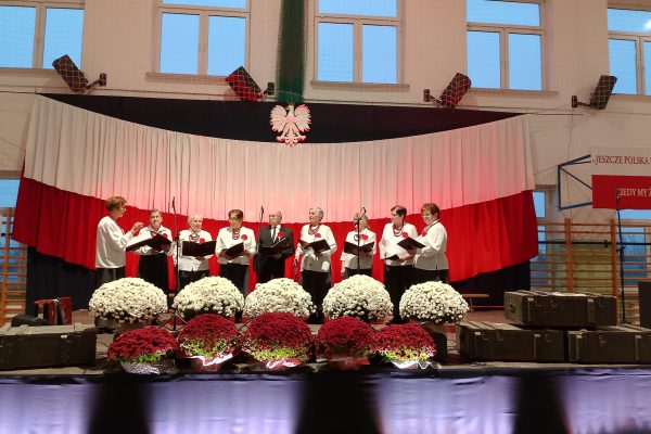 Zespół śpiewający na scenie z biało-czerwoną dekoracją i kwiatami podczas Festiwalu Pieśni Patriotycznych.
