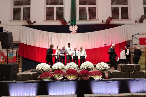 Zespół śpiewający na scenie z biało-czerwoną dekoracją i kwiatami podczas Festiwalu Pieśni Patriotycznych.