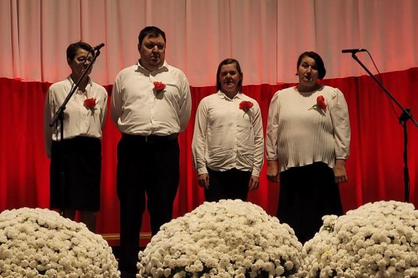 Zespół śpiewający na scenie z biało-czerwoną dekoracją i kwiatami podczas Festiwalu Pieśni Patriotycznych.