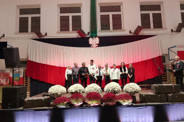 Zespół śpiewający na scenie z biało-czerwoną dekoracją i kwiatami podczas Festiwalu Pieśni Patriotycznych.
