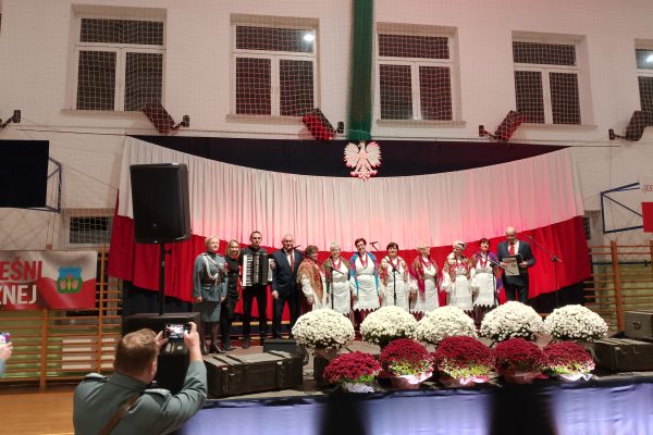 Zespół śpiewający na scenie z biało-czerwoną dekoracją i kwiatami podczas Festiwalu Pieśni Patriotycznych.