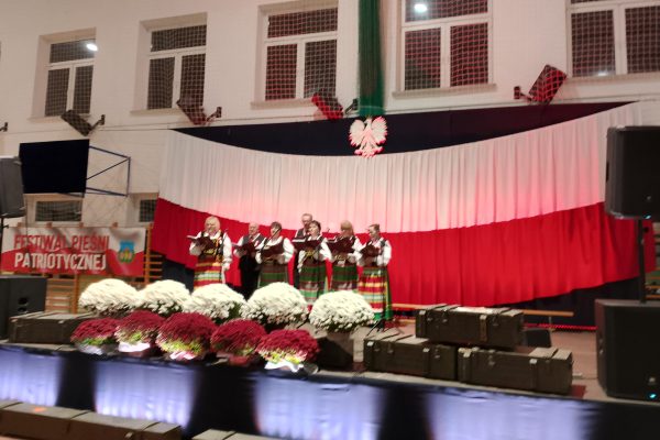 Zespół śpiewający na scenie z biało-czerwoną dekoracją i kwiatami podczas Festiwalu Pieśni Patriotycznych.