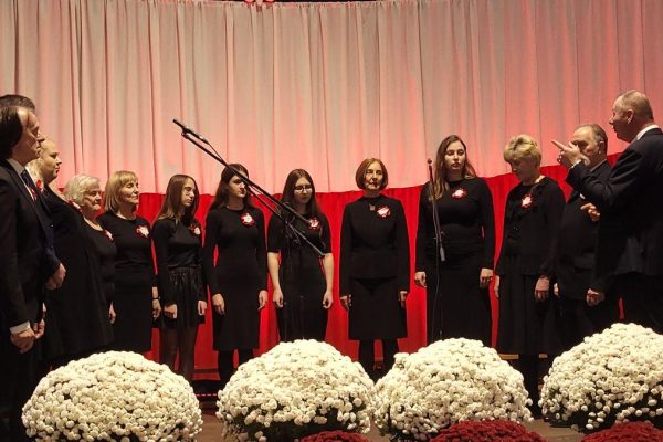 Zespół śpiewający na scenie z biało-czerwoną dekoracją i kwiatami podczas Festiwalu Pieśni Patriotycznych.