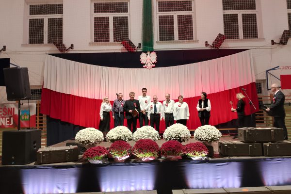 Zespół śpiewający na scenie z biało-czerwoną dekoracją i kwiatami podczas Festiwalu Pieśni Patriotycznych.