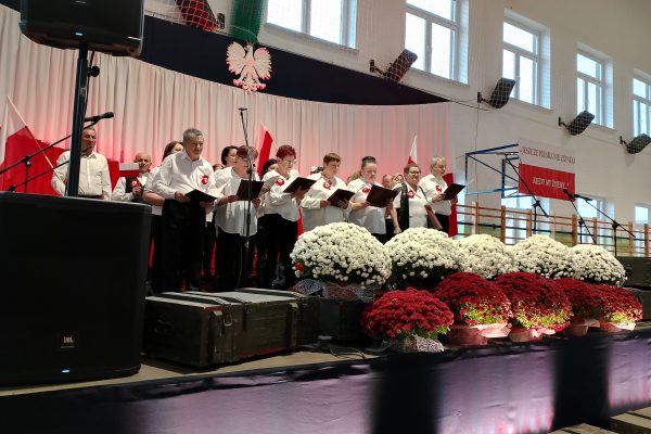 Zespół śpiewający na scenie z biało-czerwoną dekoracją i kwiatami podczas Festiwalu Pieśni Patriotycznych.