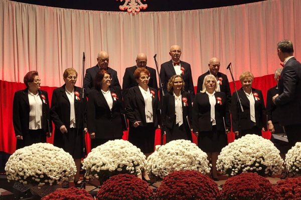 Zespół śpiewający na scenie z biało-czerwoną dekoracją i kwiatami podczas Festiwalu Pieśni Patriotycznych.