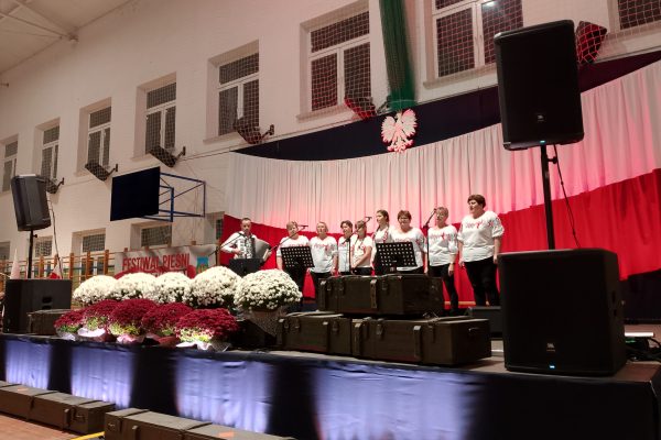 Zespół śpiewający na scenie z biało-czerwoną dekoracją i kwiatami podczas Festiwalu Pieśni Patriotycznych.