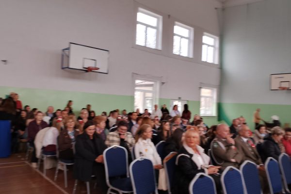 Publiczność i osoby przy stole jury podczas Festiwalu Pieśni Patriotycznych w sali sportowej.