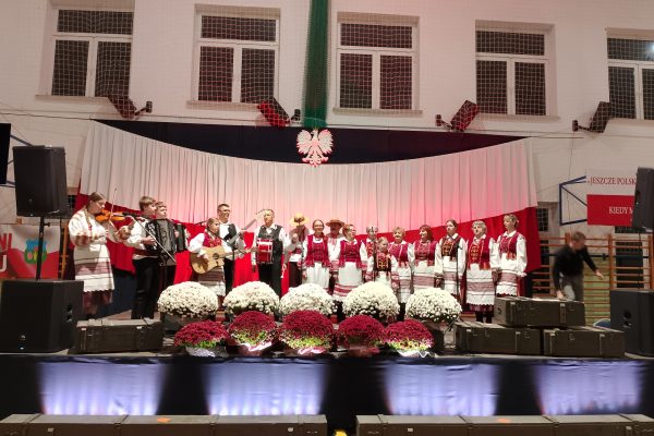 Zespół śpiewający na scenie z biało-czerwoną dekoracją i kwiatami podczas Festiwalu Pieśni Patriotycznych.