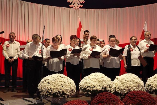 Zespół śpiewający na scenie z biało-czerwoną dekoracją i kwiatami podczas Festiwalu Pieśni Patriotycznych.