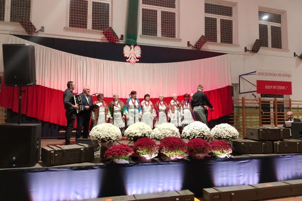 Zespół śpiewający na scenie z biało-czerwoną dekoracją i kwiatami podczas Festiwalu Pieśni Patriotycznych.