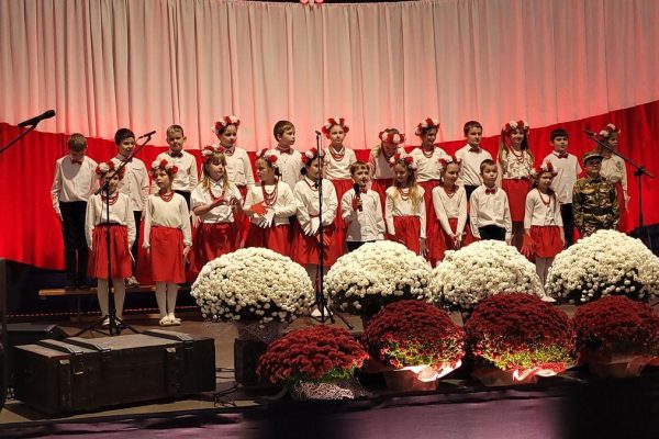 Zespół śpiewający na scenie z biało-czerwoną dekoracją i kwiatami podczas Festiwalu Pieśni Patriotycznych.