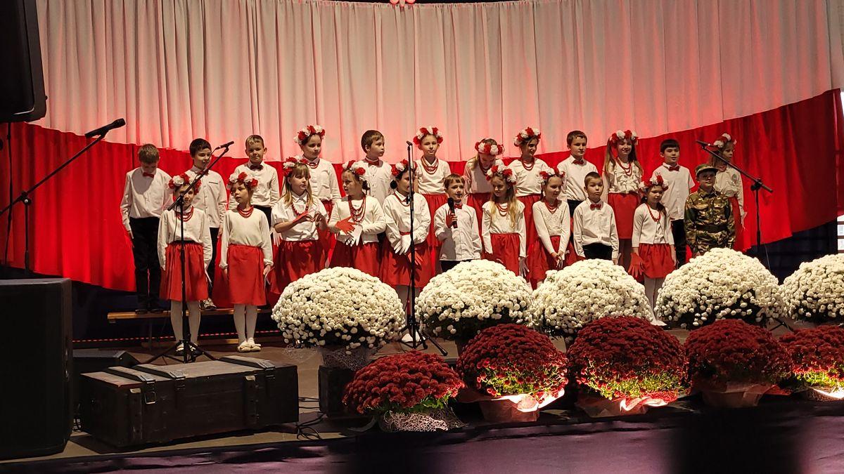 Zespół śpiewający na scenie z biało-czerwoną dekoracją i kwiatami podczas Festiwalu Pieśni Patriotycznych.